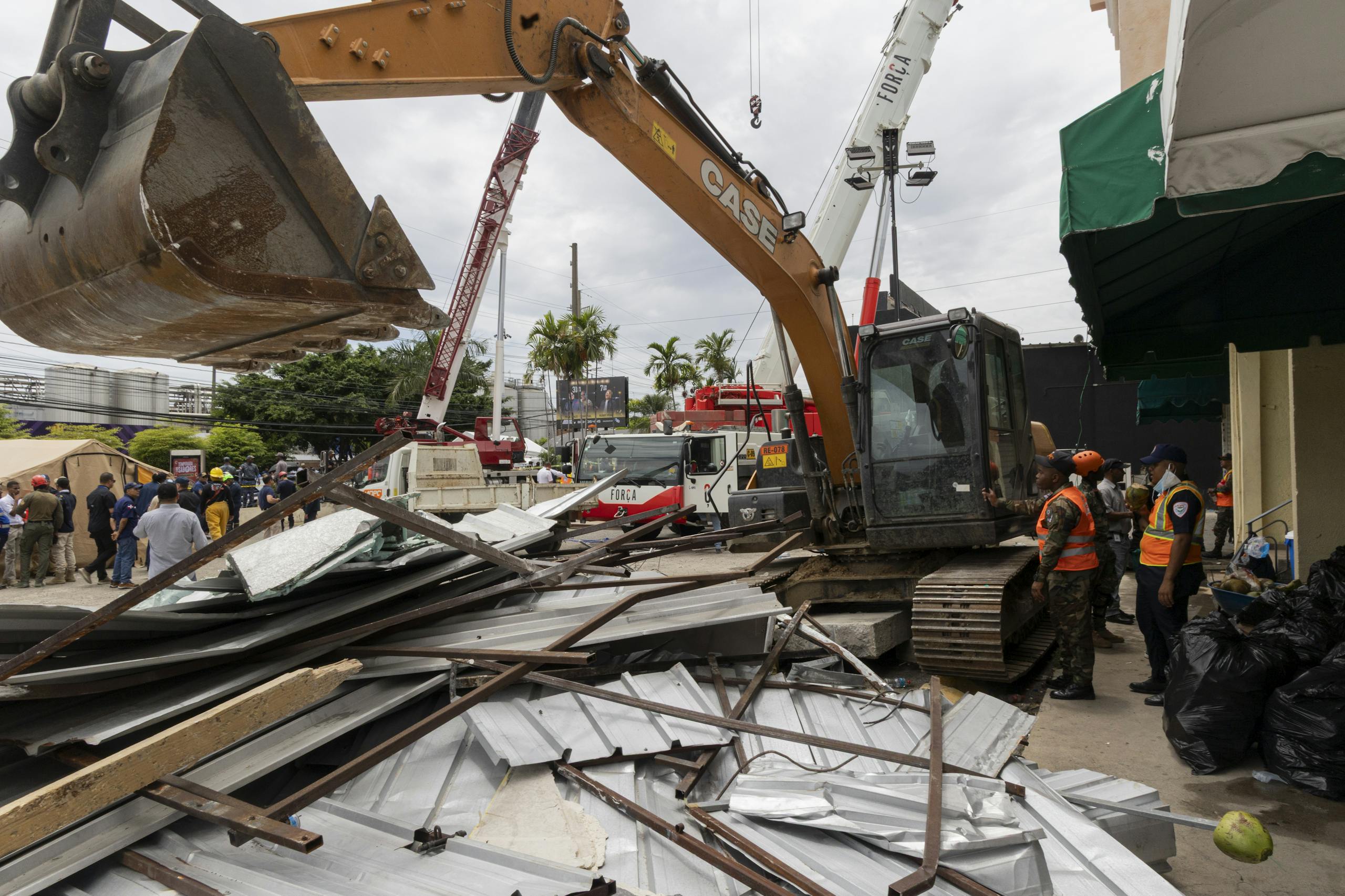 Excavator clearing debris after emergency in urban area with workers and rescue vehicles.
